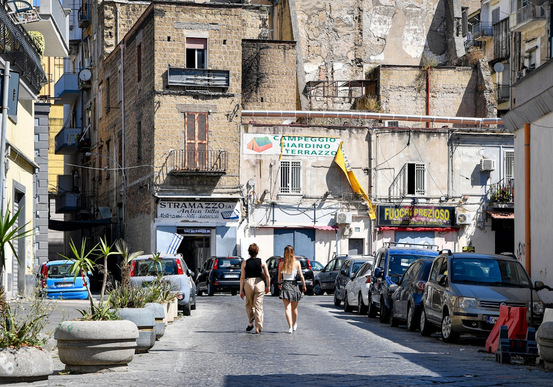 Two women walk down a narrow street lined with cars.