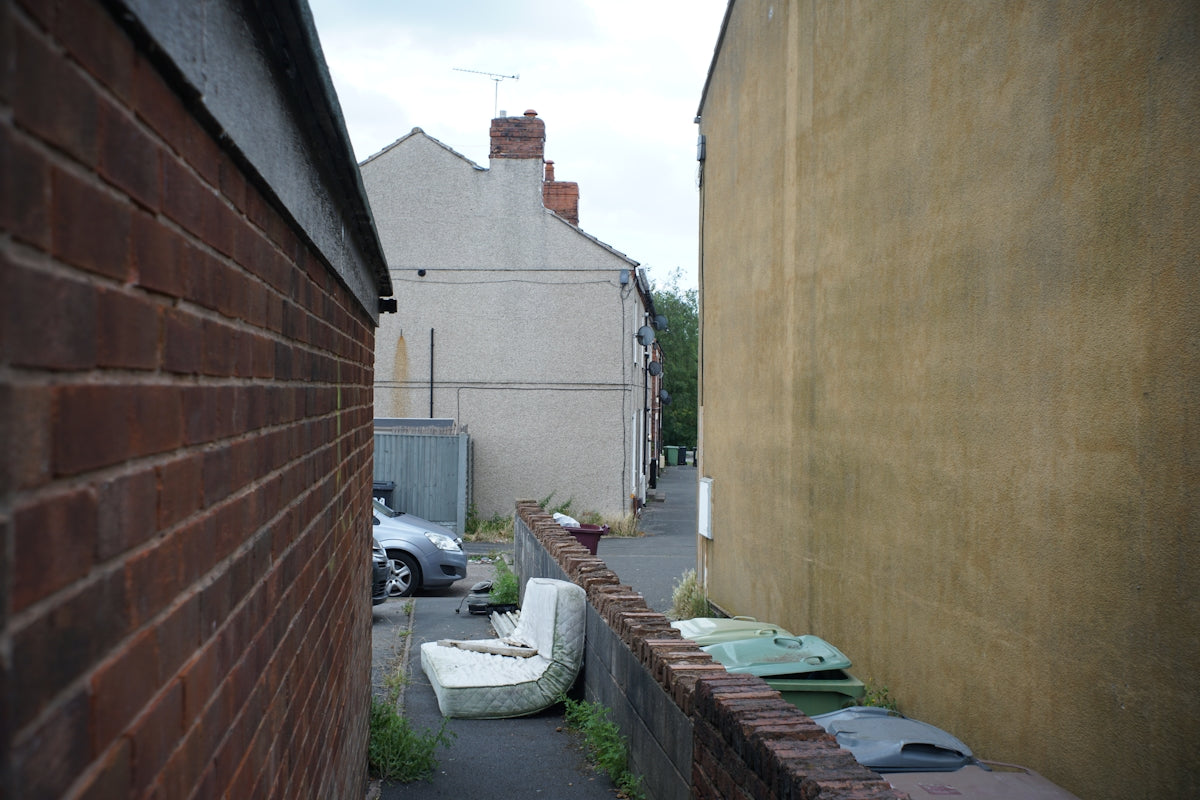 Narrow alleyway between brick and yellow buildings with discarded items.