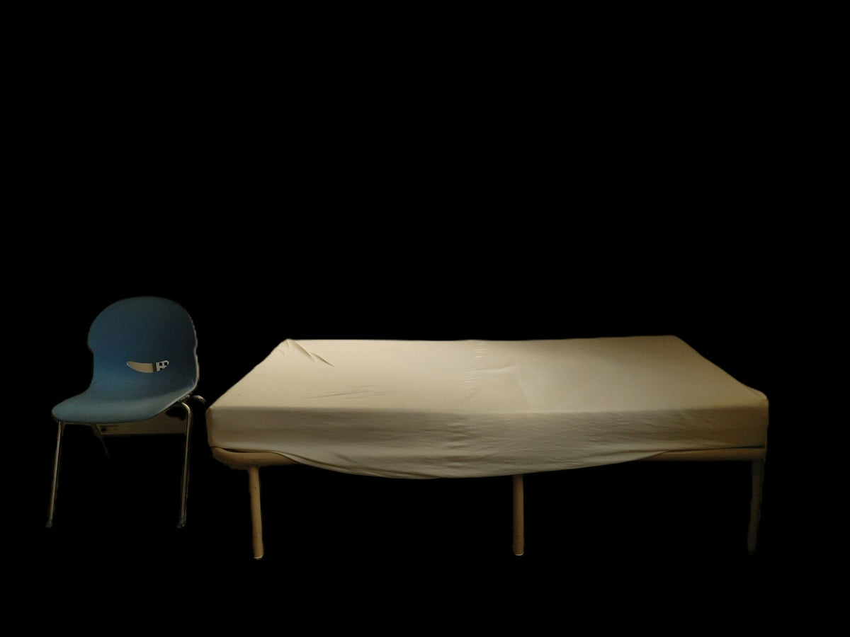 A blue plastic chair beside a bed with a white sheet against black