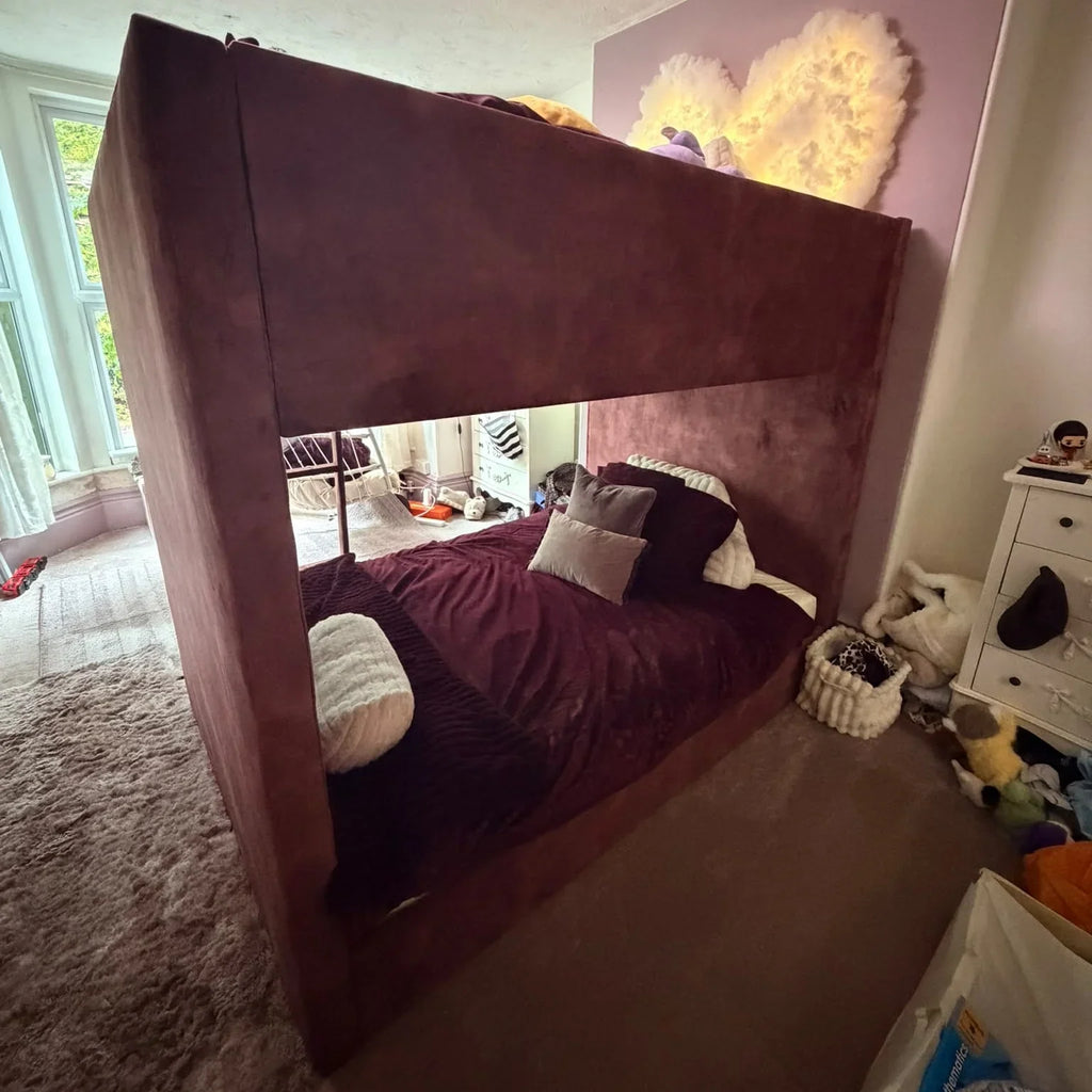 A large upholstered bunk bed frame in a Ox Blood Coniston fabric, shown in a real bedroom with bedding, pillows, a window, rug, and storage items visible.