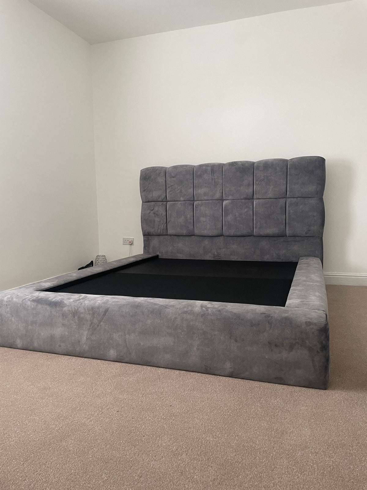 A gray upholstered platform bed frame with a tall, square, tufted headboard, shown in a minimalist room with white walls and beige carpet.
