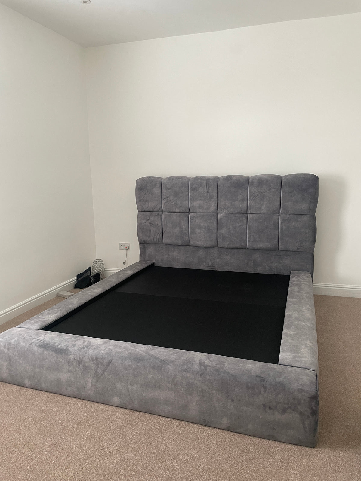 A gray upholstered platform bed frame with a high tufted headboard, shown in a bedroom setting with beige carpet and white walls.