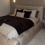 A modern upholstered bed in a bedroom setting, featuring a tufted cream Plush headboard and matching footboard, dressed with white bedding, a dark brown throw blanket, and black accent pillows.