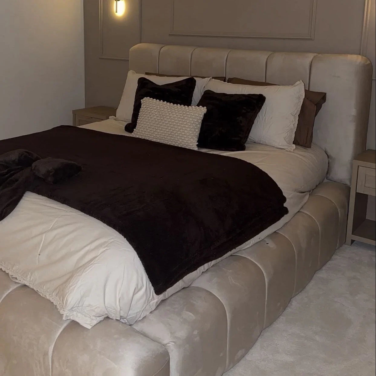 A modern upholstered bed in a bedroom setting, featuring a tufted cream Plush headboard and matching footboard, dressed with white bedding, a dark brown throw blanket, and black accent pillows.